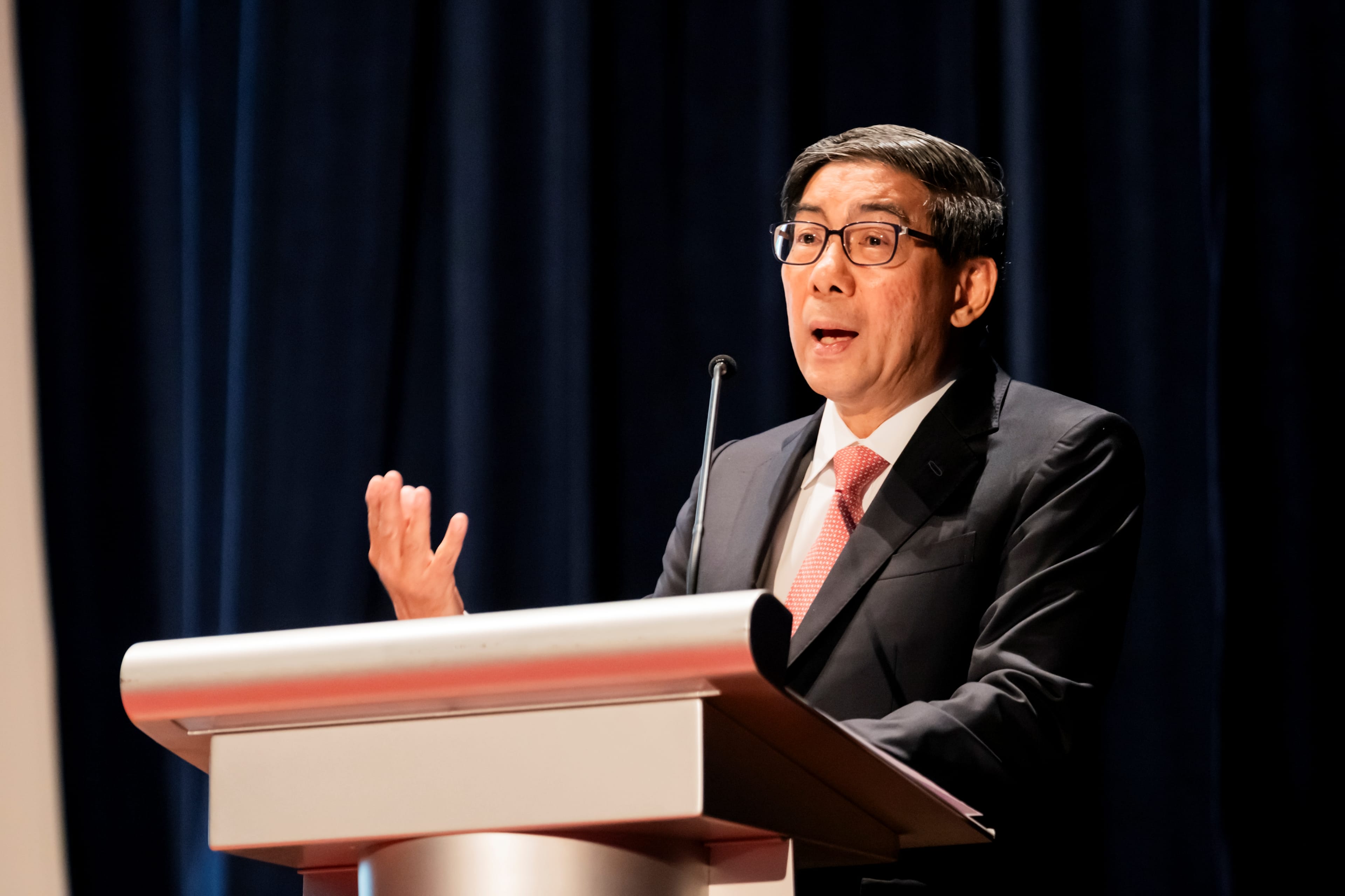 Guest of Honour, Dr Yeo Li Pheow, donning a dark suit, and a pink tie speaking at a podium with a microphone, gesturing with his right hand against a dark blue backdrop.